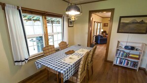 Un comedor con mantel a cuadros, sillas de madera, una araña y una estantería llena de libros.