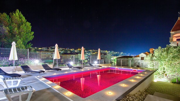 A swimming pool with lounge chairs and umbrellas, surrounded by a fence and a view of a cityscape at night.