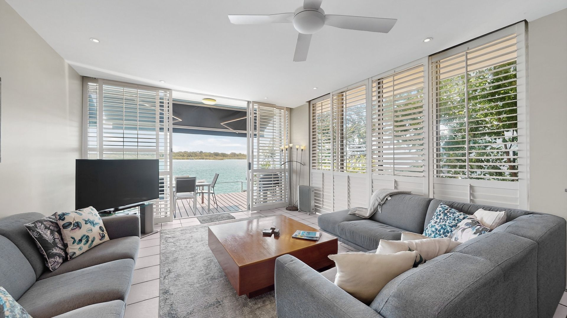 A modern living room with a large TV, grey sofas, a coffee table, and large windows with blinds.