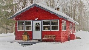 Una pequeña cabaña roja con puerta y ventanas blancas, rodeada de nieve y árboles.