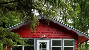 Una cabaña roja con puerta blanca, ventanas amplias y un porche pequeño con un banco.