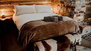 A cozy bedroom with a wooden headboard, a stone wall, and a plush bed with a brown blanket and a patterned throw.