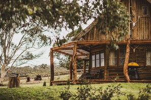 A rustic wooden cabin with a porch, a grill, and a tree in the foreground. - Kallarroo Cottage - Rustic Log Cabin Retreat (Greenlands)