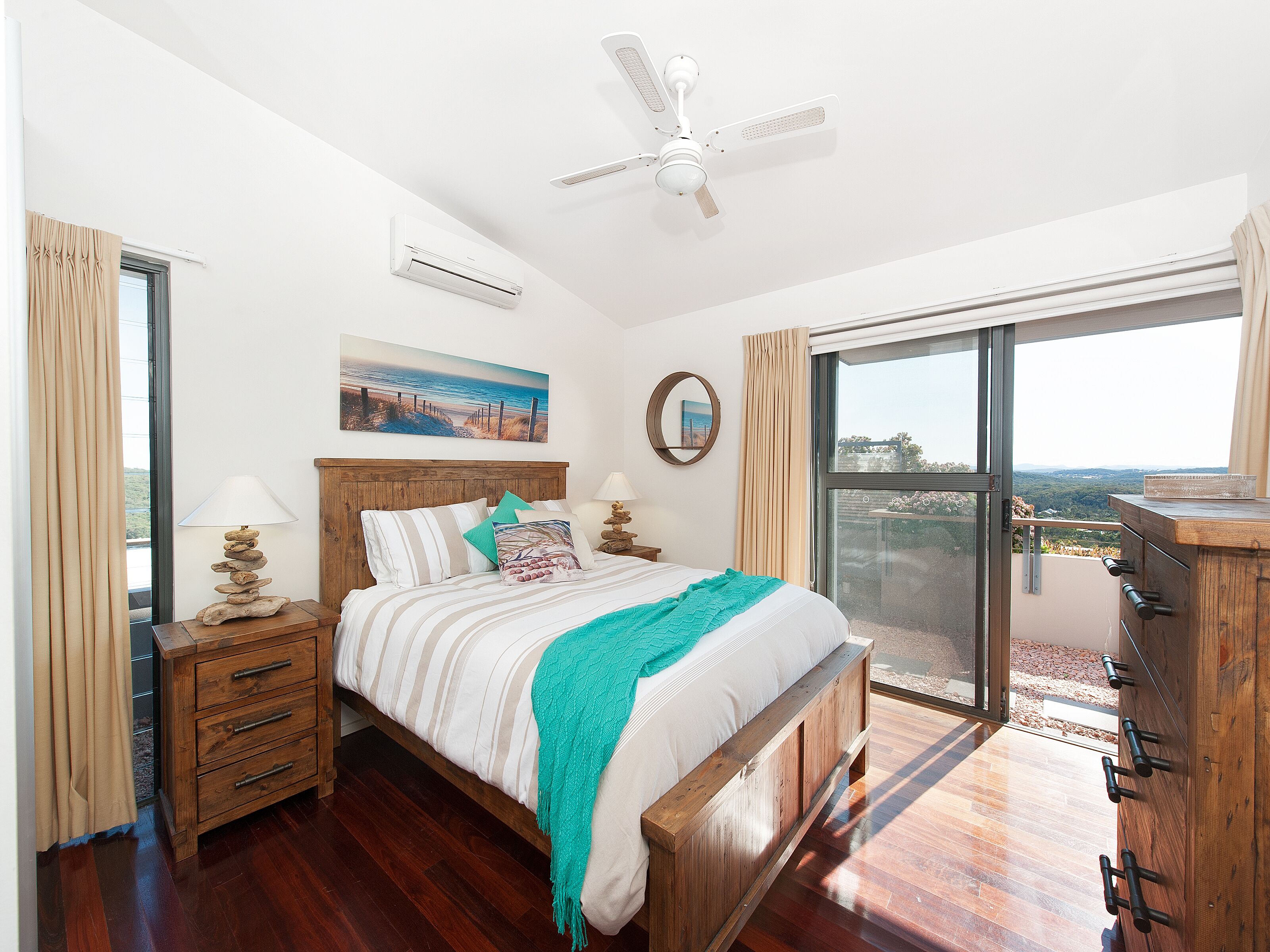 A bedroom with a bed, wooden headboard, bedside tables, a ceiling fan, and a large window with a view.