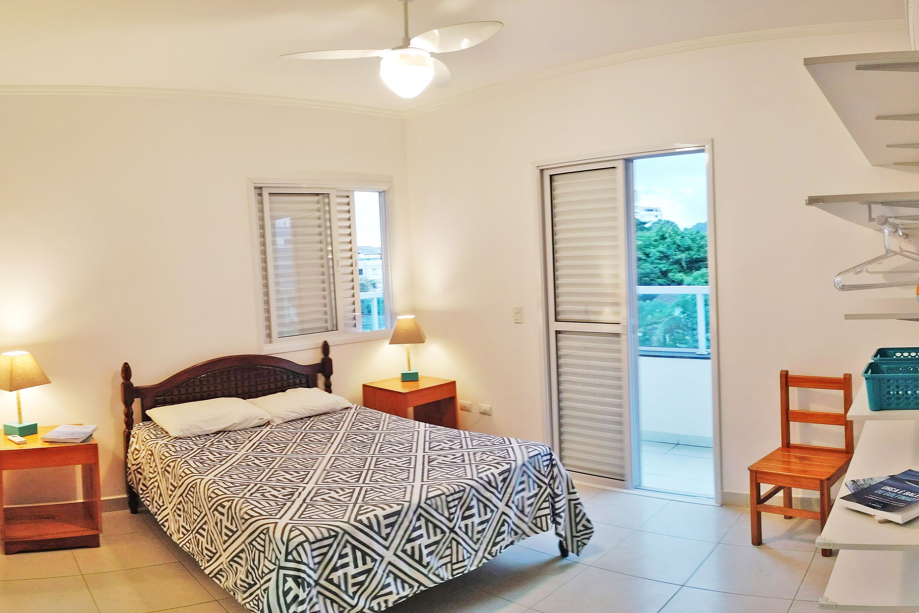Une chambre à coucher comprenant un lit, deux tables de chevet, un ventilateur de plafond, une fenêtre avec stores et une porte-fenêtre donnant sur un balcon.