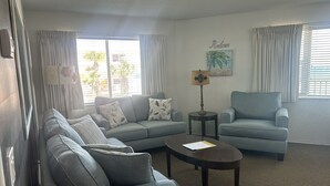 A living room with a grey sofa, a blue armchair, a coffee table, and a lamp. There are curtains on the windows, and a painting with the word "Relax" on the wall. - Spacious 3 Bedrm/2 Bth OceanFront Apt w/Large Balcony Overlooking Pool. Sleeps 9 (Daytona Beach Shores)