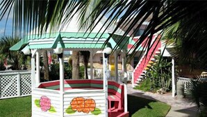 A children's play structure with a green roof and pink accents, surrounded by palm trees and a white fence. - Spacious 3 Bedrm/2 Bth OceanFront Apt w/Large Balcony Overlooking Pool. Sleeps 9 (Daytona Beach Shores)