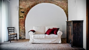A white sofa with red cushions, a wooden chair, and a dark wooden cabinet in a room with a brick wall and white ceiling.