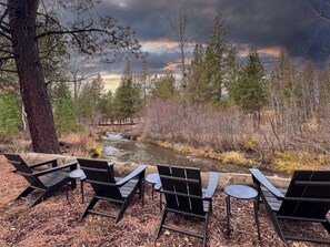 A riverside seating area with chairs and tables, surrounded by trees and a wooden bridge. - Sisters, OR Lodge – Pickleball, Private Creek, Hot Tub & Mt Views on 8.5 Acres (Sisters)
