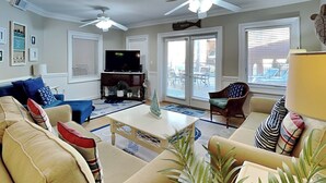 A living room with a sofa, armchairs, a coffee table, and a TV. There are ceiling fans, a window with blinds, and a door.