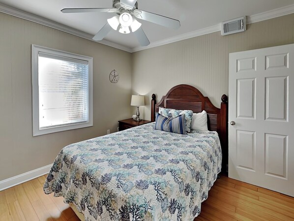 Une chambre à coucher comprenant un lit, un ventilateur de plafond, une fenêtre avec des stores, une table de chevet avec une lampe et une porte.