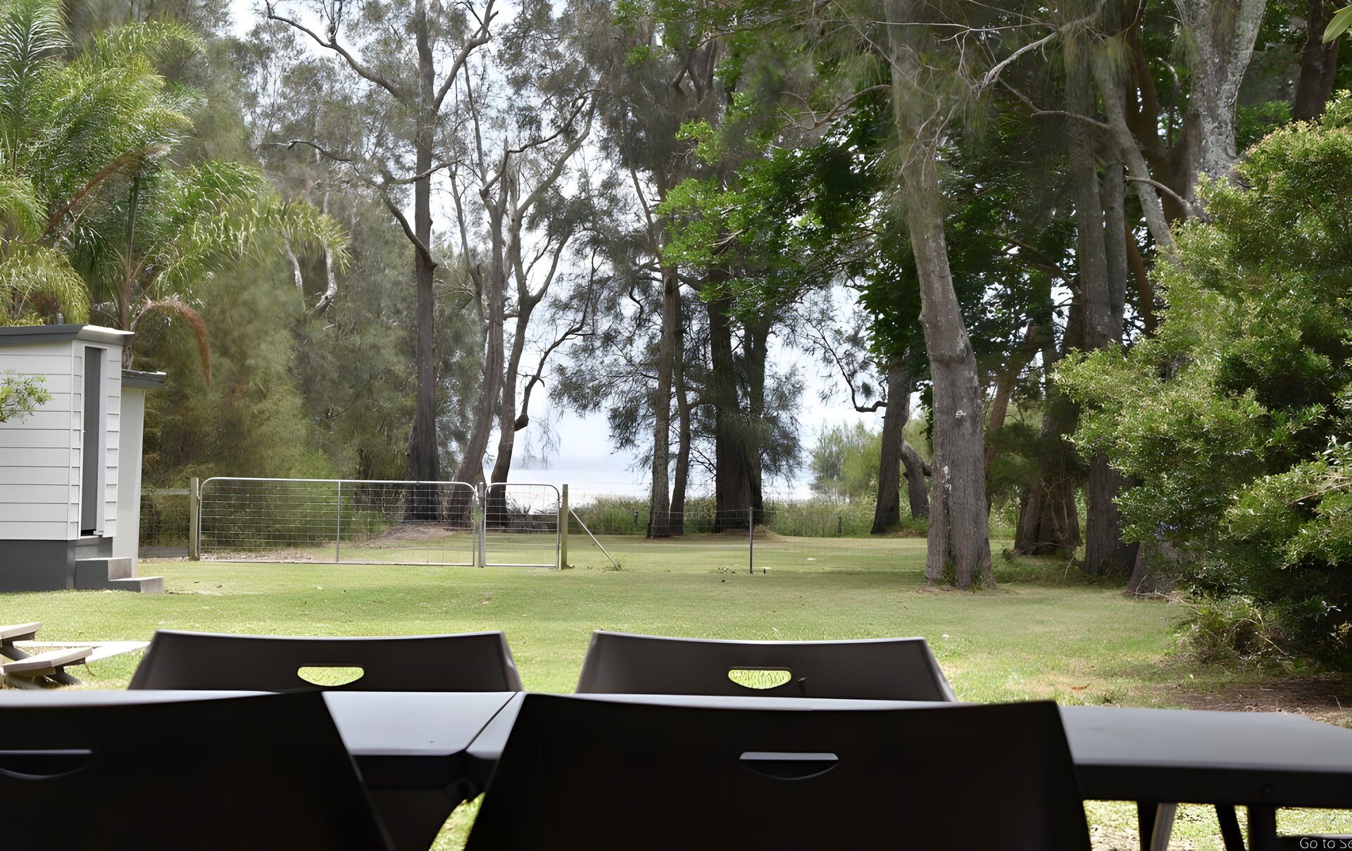 Een terras met tafels en stoelen, omringd door bomen en een omheind sportveld.