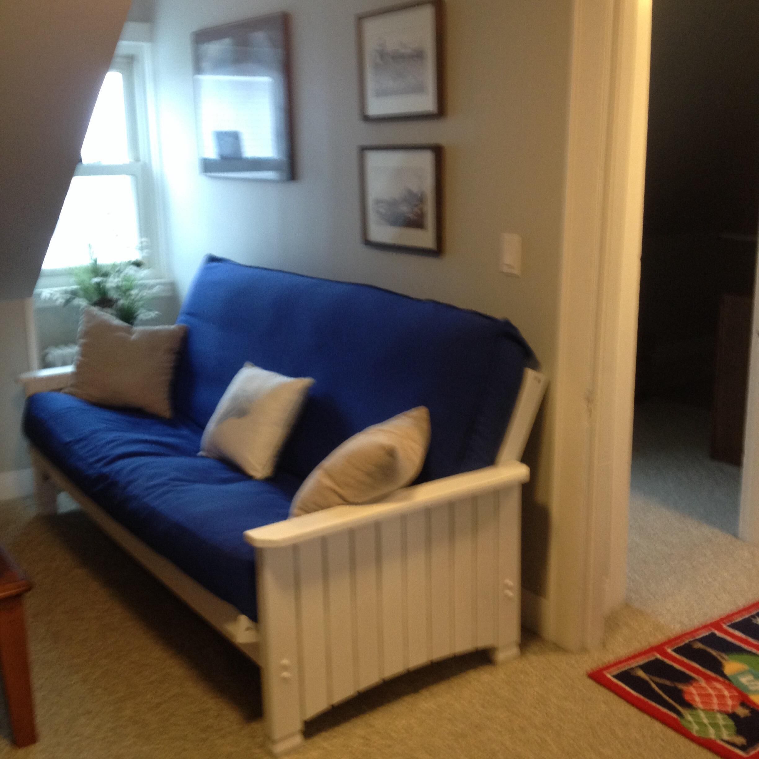 A blue sofa with white cushions in a room with a radiator, a window with a view, and framed pictures on the wall.