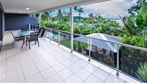 A spacious balcony with a glass railing, a dining area with chairs and a table, and a view of lush greenery and mountains in the distance. - Blue Water Views 9, Seaview Apartment on Hamilton Island (Hamilton Island)