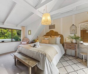 A bedroom with a large bed, a bench, a wooden headboard, a hanging light fixture, a small table with a plant, and a view of greenery outside.
