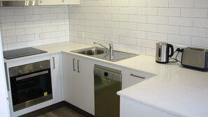 A compact kitchen with white cabinets, a stainless steel sink, a dishwasher, a toaster, and a built-in oven. - API Leisure & Lifestyle Battery Point (Battery Point)