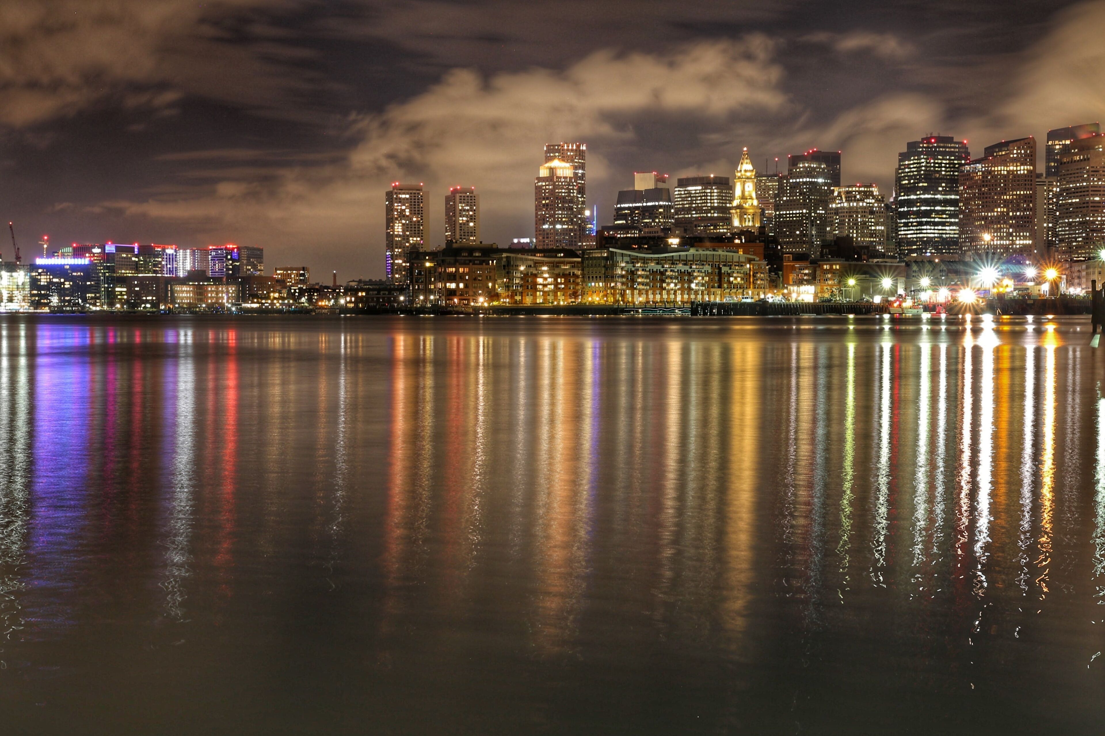 A nighttime city skyline with illuminated buildings reflected in the water.