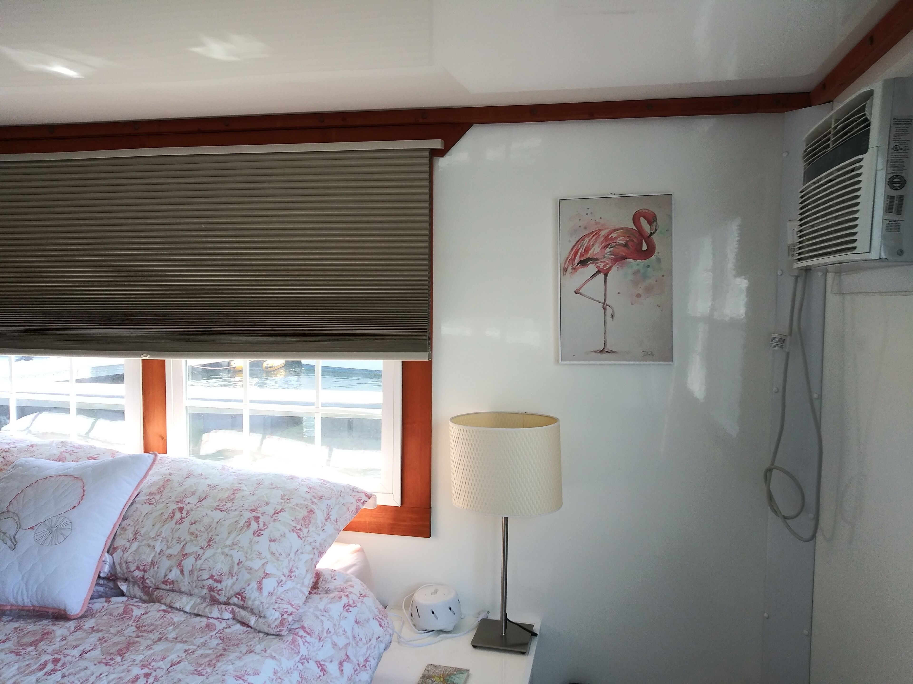 A bedroom with a bed, a window with blinds, a wall-mounted air conditioner, and a painting of a flamingo.
