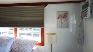 A bedroom with a bed, a window with blinds, a wall-mounted air conditioner, and a painting of a flamingo.