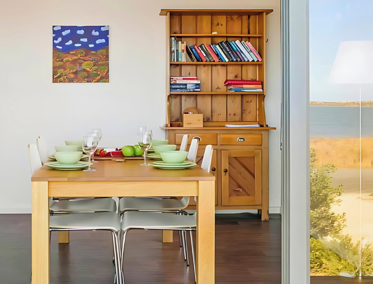Un comedor con una mesa de madera dispuesta para dos personas, una estantería con libros y un cuadro en la pared.