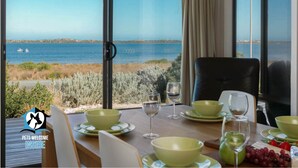 A dining table with green bowls and wine glasses, a chair, and a view of the sea through large windows. - Absolute Waterfront Views + Kayaks (Noonameena)