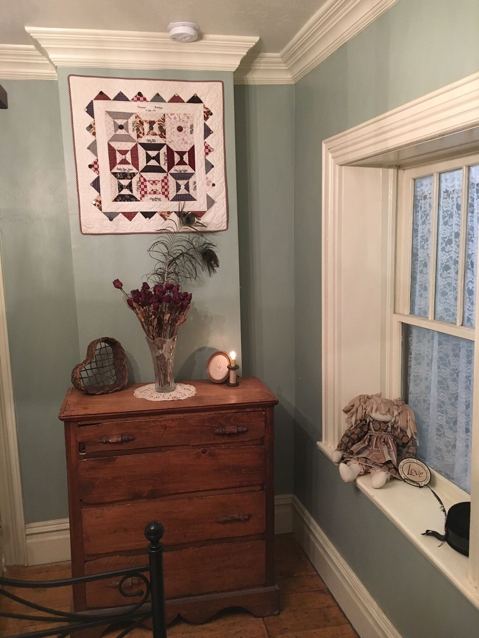 Une pièce avec une commode en bois, une courtepointe accrochée au mur, un vase rempli de fleurs et une fenêtre ornée d’un rideau en dentelle.
