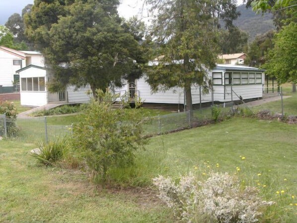 A white mobile home with a large tree in front, surrounded by a fenced yard and a grassy area with wildflowers. - Kinder Cottage Talbingo. (Talbingo)