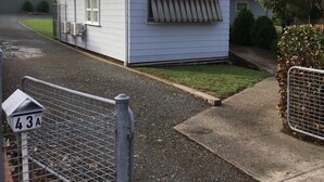 A small, single-story mobile home with a window and a door, a small porch area, and a fenced driveway.