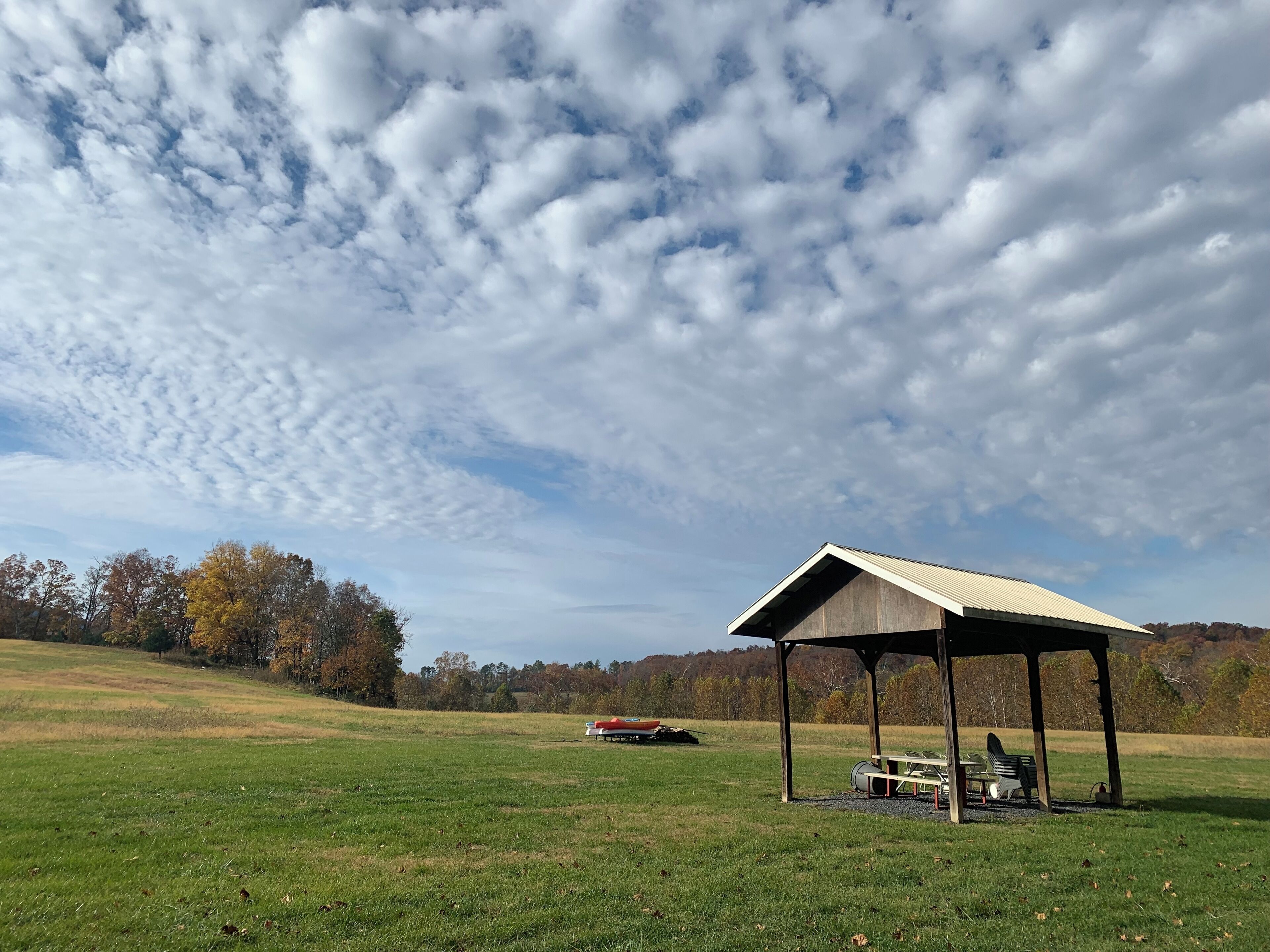 A gazebo in a grassy field with a picnic table and benches, surrounded by trees and a clear sky.