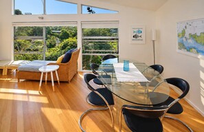 Une pièce moderne avec une table à manger en verre, des chaises noires, un canapé en osier et de grandes fenêtres donnant sur un paysage verdoyant.