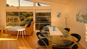 A modern room with a glass dining table, black chairs, a wicker sofa, and large windows offering a view of greenery. - The Palms - Sea Views near Sandy Bay (Sandy Bay (Waiheke))