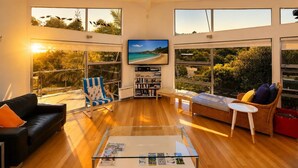 A spacious living room with a large TV, wooden flooring, and large windows offering a view of the beach. - The Palms - Sea Views near Sandy Bay (Sandy Bay (Waiheke))