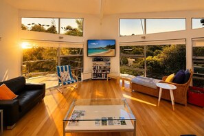 A spacious living room with a large TV, wooden flooring, and large windows offering a view of the beach.