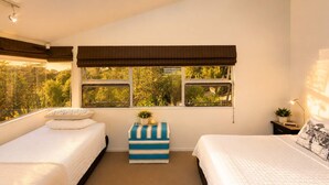A bedroom with two beds, a window with a view of greenery, a bedside table with a lamp, and a small stool with a plant. - The Palms - Sea Views near Sandy Bay (Sandy Bay (Waiheke))