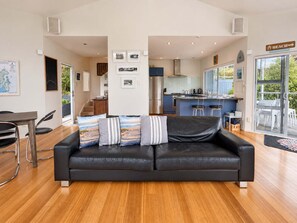 A modern living room with a black leather sofa, wooden flooring, and a kitchen in the background.