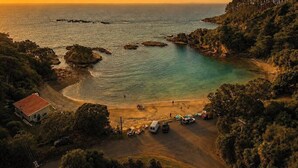 A coastal area with a beach, a small building, and parked vehicles. - The Palms - Sea Views near Sandy Bay (Sandy Bay (Waiheke))