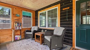 A cozy log cabin interior with wicker furniture, a bookshelf, and a large window. - Rangeley Manor 2 | Great Cabin with Shared Access to Rangeley Lake. (Rangeley)