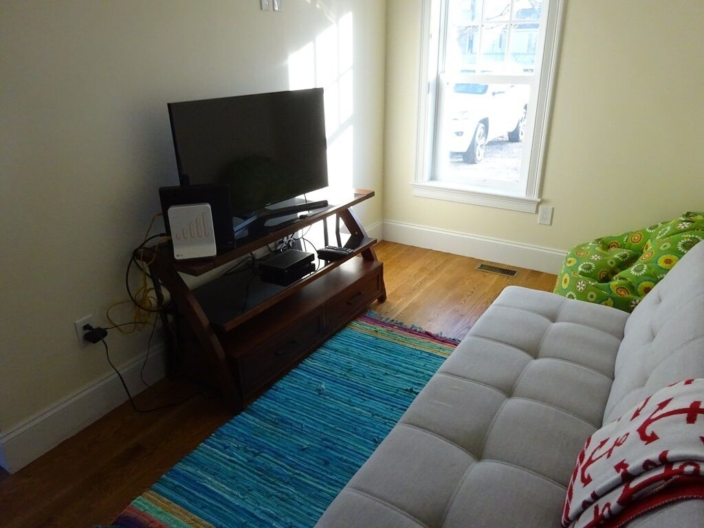 Uma sala de estar com uma televisão de tela plana sobre um suporte de madeira, um sofá branco e um tapete colorido.