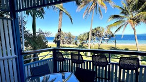 Un balcón con mesa y sillas de vidrio, con vistas a una playa con palmeras y el océano.