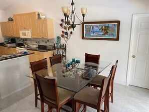 A dining area with a glass table and wooden chairs, a kitchen with wooden cabinets, a microwave, and a painting on the wall.