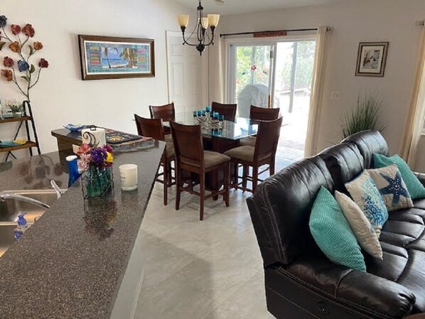 A dining area with a glass table and chairs, a kitchen counter with a sink, and a leather couch with decorative pillows.