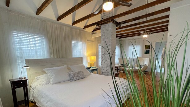 A bedroom with a large bed, wooden beams, a white brick column, and a view of the living area. - Magical Little Home in Historic District • Parking  • Rustic Luxury  • Outdoor.. (Marblehead)