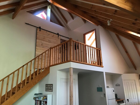 A loft-style room with a wooden staircase, a lofted seating area, and a kitchen with a refrigerator and sink.