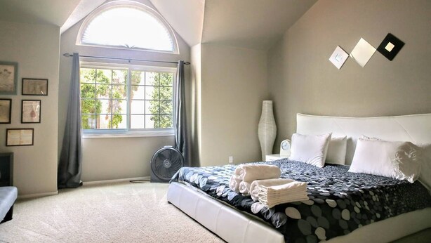 A bedroom with a large bed, a window with curtains, a bedside table with a lamp, and framed pictures on the wall.