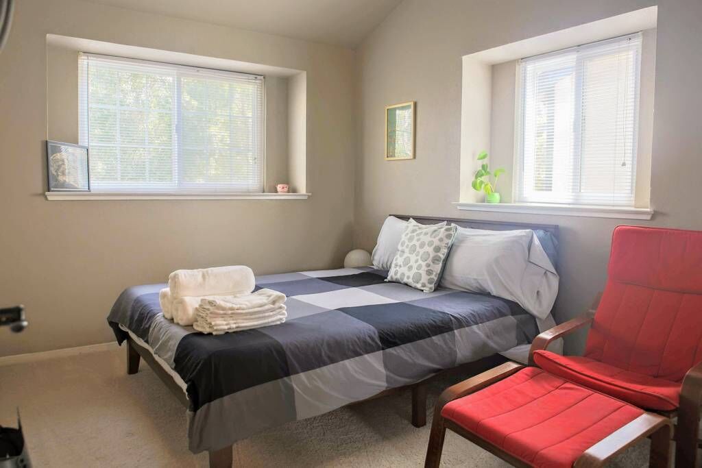 Ein Schlafzimmer mit einem Bett, zwei Fenstern, einem Sessel und einer kleinen Pflanze.