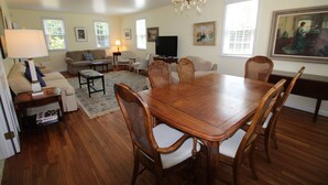 A spacious living room with a wooden dining table, comfortable seating, a television, and artwork on the walls.