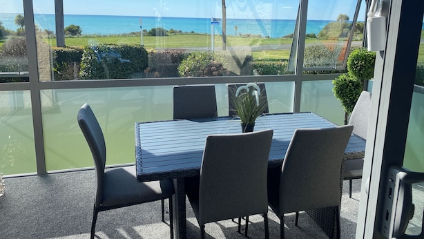 Un balcón acristalado con una mesa y sillas, con vista a un mar azul cristalino y un paisaje verde.