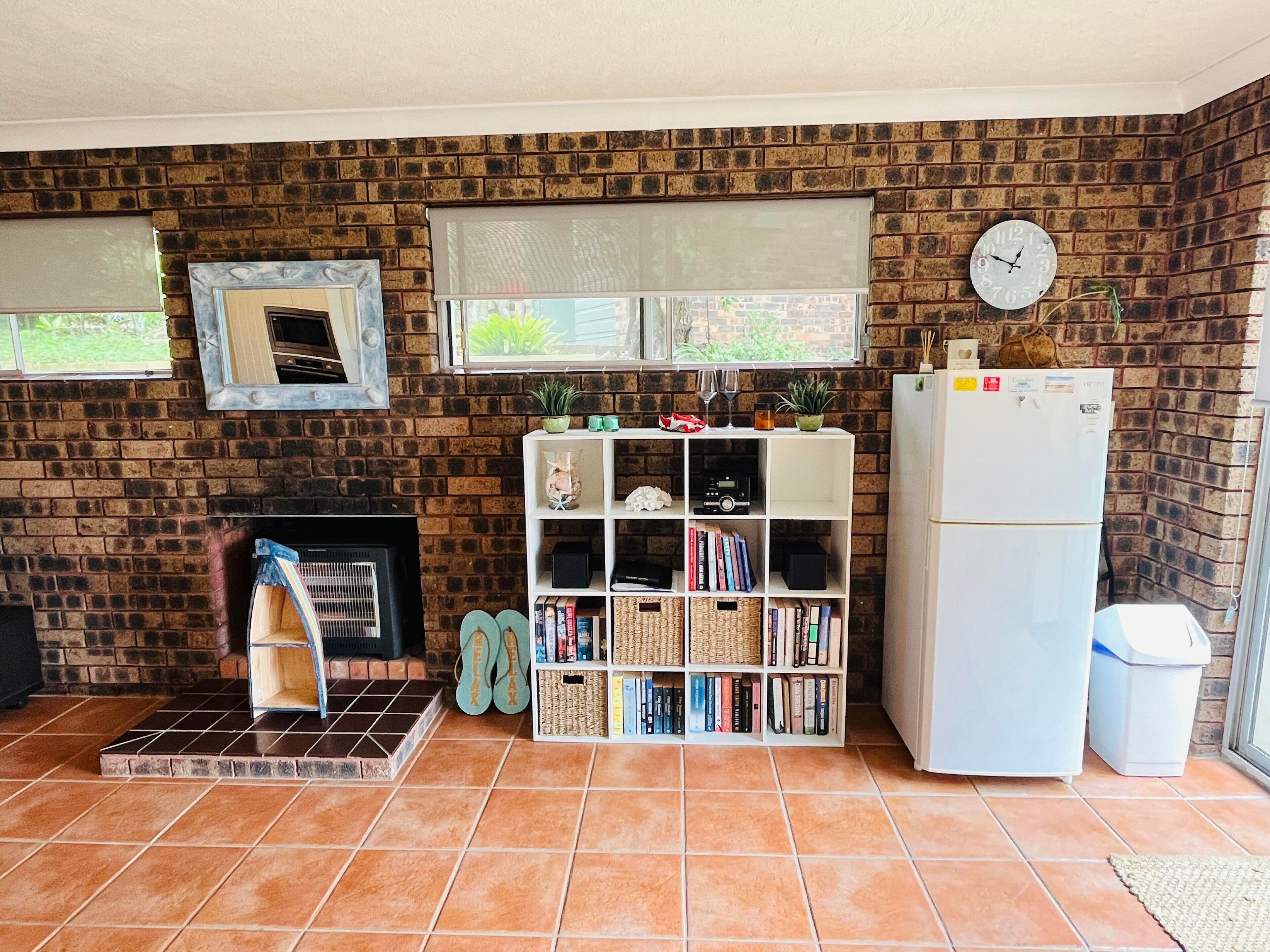 A living room with a brick wall, a fireplace, a bookshelf, a clock, and a refrigerator.