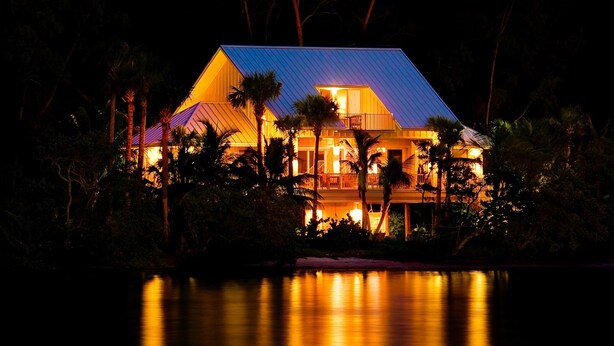 A large, two-story house with a metal roof, surrounded by palm trees and reflecting in a body of water. - Private Island Retreat with Private Beach,  Dock, Palm Trees & Glorious Sunsets (Longboat Key)