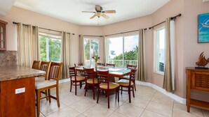 A dining area with a large table and chairs, a ceiling fan, and large windows with curtains. - Key Largo Sunset Estate (Key Largo)